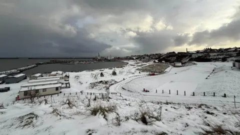 A wintry scene at Peterhead - snow covers the ground and roads, with the harbour and sea visible in the distance