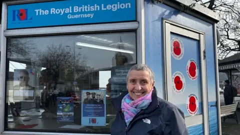 BBC A smiling woman in a blue jacket in front of a hexagonal kiosk, which is decorated with poppy stickers and a Royal British Legion Guernsey sign.