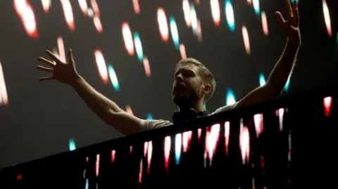Calvin Harris stands on a dark stage behind DJ decks. The stage is lit up by stage lights.