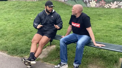 Bristol and England rugby player Ellis Genge and actor Joe Sims sit together on a bench in Redcatch Park and talk. They are looking at each other and Genge is indicating something with his hands.