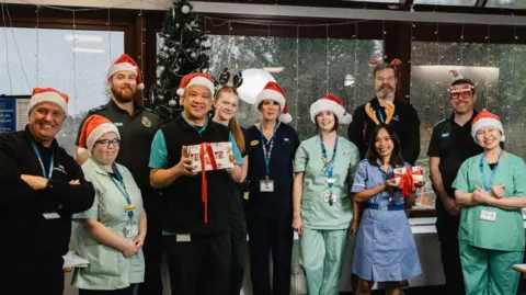 Torbay and South Devon NHS Charity A group of hospital staff in green and blue medical uniforms are wearing read and white Santa hats and antlers.