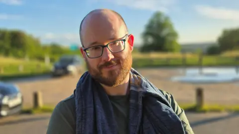 Paul Moseley/BBC A close-up outdoor picture of Stephen Robinson. He is standing in a car park near an open green space. Vehicles and wooden posts are visible behind him, with warm sunlight and a shallow depth of field.