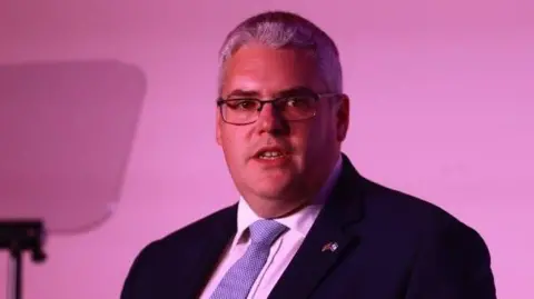 PA Gavin Robinson pictured in a suit in front of a pink background