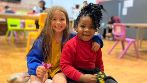 A child with long, light brown hair, and a yellow and blue top, has her arm around another child with black curly hair. They're both smiling and sitting on a wooden floor inside a holiday club.