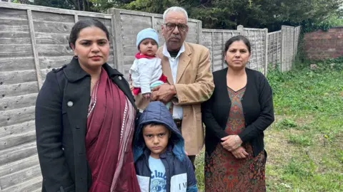 Kate Bradbrook/BBC Five people, including two children, stand together outdoors by a wooden fence.
