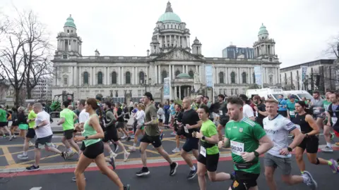 Pacemaker Press A large group of people are running together. Many are wearing green. Belfast City Hall is in the background. The sky is overcast. 