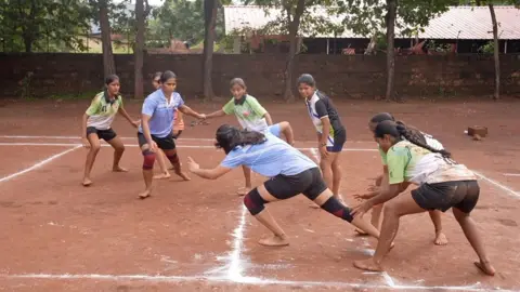 Action shot of seven girls playing Kabdi. They are purple, green and black sports shirts, shorts and are barefoot. The pitch is bare earth with white lines marked on it. 