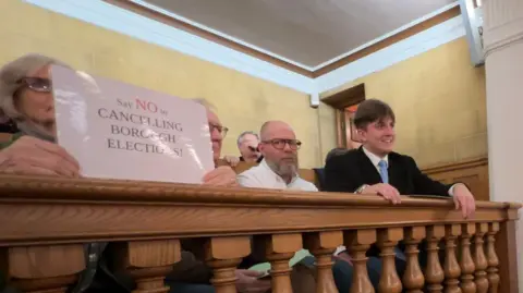 George Finch sits smiling while leaning on a wooden barrier. A woman to the left of the image holds a sign which reads "say no to cancelling borough elections".