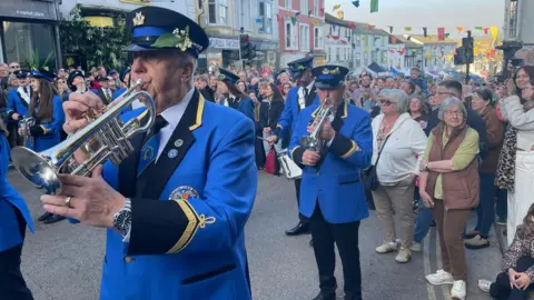Two members of the band are playing trumpets. They are wearing smart bright blue uninforms, complete with hats which have sprigs of Lily of the Valley tucked into the hat band. The street is full of crowds.