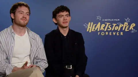 Actors Kit Connor and Joe Locke in front of a banner for Netflix's Heartstopper Forever.