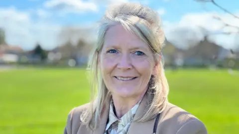 Emma Coles as a grown woman standing in a green field as the sun is shining. She has shoulder-length blonde hair and is smiling. She is wearing a beige jacket and a beige and white shirt.
