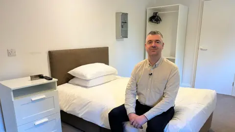 Nick Dunkley, a man with short grey hair, is wearing a beige shirt, black trousers and brown shoes. He's sitting on a bed in a resident's room at Banbury Lodge. The room is painted white, with white sheets and pillows on the bed and white furniture. 