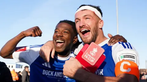 Macclesfield FC goalscorers Paul Dawson and Isaac Buckley-Ricketts, wearing the club's blue-and-white shirts, with their arms around each other as they celebrate on the pitch after the final whistle.