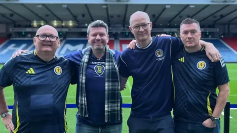 BBC Cuatro fanáticos masculinos de Escocia, todos con camisetas azul marino de Escocia, están del brazo con el campo y los asientos detrás de ellos en Hampden Park.