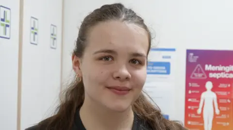 Jamie Niblock/BBC A young woman smiles at the camera. She is standing in a pharmacy room. She has brown hair that has been tied back, some of which hangs over her shoulders. She is wearing a black top and has gold hoop earrings.