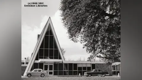 Swindon Borough Council A black and white photo of a building with a very sharp, pointed roof on the left and a flat one-storied area to the right. There are two cars parked at the front and there is a large tree on the right of the picture and a title saying 'Swindon Libraries c1966 in the top left-hand corner
