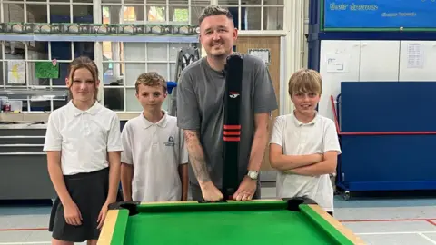 Three children stand alongside Kyren Wilson in a school hall, next to a mini snooker table. 