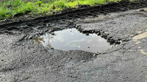 Aa large pothole filled with muddy water on what looks like a worn tarmac road.