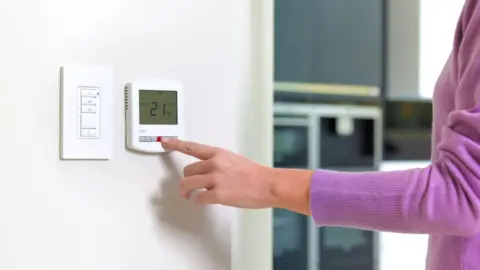 A woman in a purple jumper presses a button on a white thermostat. The thermostat, which sits on a white wall, reads the temperature 21 degrees Celsius. Beside the thermostat, on the wall, is a white multi-button panel.