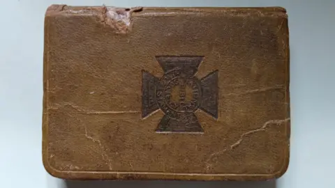 Macfarlane family A brown leather bible with a military cross emblem on the front cover. On the left hand side of the bible, close to the spine, the leather has been broken with some white material seen beneath