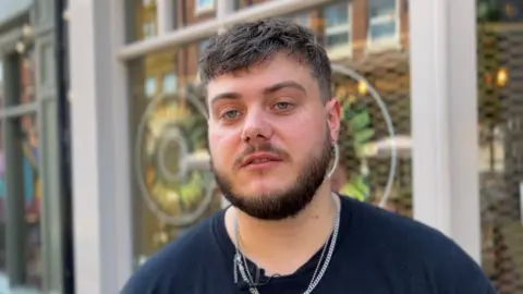 A picture of Cameron Bedford, who has brown hair and a brown beard standing in from a a bar on Kirkgate in Leeds, there are windows behind him.