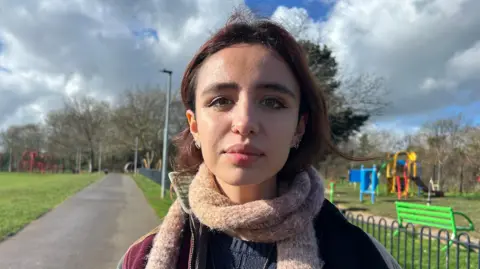 A young woman with dark brown hair and a brown knitted scarf