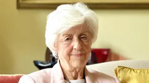 Lady Diana Brittan pictured sitting on a cream sofa with one golden cushion, and another red cushion with golden medieval tapestry print, with yellow walls behind her and the golden frame of an artwork just visible. She is wearing a light pink linen jacket, white blouse. She is in her 80s and has white hair, and is pictured smiling slightly.