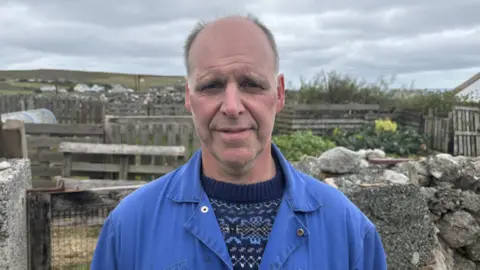 John Arthur Poleson standing outside in a croft. He is wearing a patterned jumper covered by blue overalls. There are stone walls and gates behind him and hills in the distance. He is bald and has some facial hair.