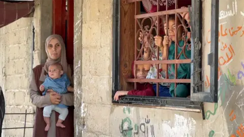 Nawara, who is wearing a brown headscarf and matching tunic, holds baby Rahma in her arms in the doorway of their home. The brick looks damaged and there is graffiti. Looking out of the window, which is open and covered by a metal grill, are some of the other children. Malak is on the left, the patterned t-shirt of another child can be seen in the middle, and the child on the right is kneeling on the window sill and holding onto the bars.