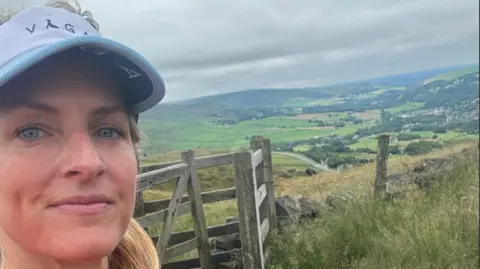 Mel Sykes A woman with blonde hair standing in front of a gate overlooking hills an fields 