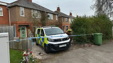 A police van is parked on the drive of a house. Police tape stretches across the drive from a green bin to a fence panel. Some flowers have been laid to the left of the drive, by the fence panel. The front door of the house is green with red brick around the lower level of the house it and brown render above.
