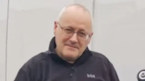 A middle aged bald man with glasses standing in front of a white van wearing a blue jumper