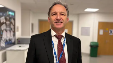 A man in a black jacket and burgundy spotted tie with a blue lanyard in a school corridor. It is a head and shoulders shot.
