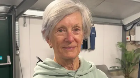 Tom Jackson/BBC Celia Duff is smiling at the camera and wears a light green hoodie and small gold hooped earrings. She has light grey short hair and a side fringe.