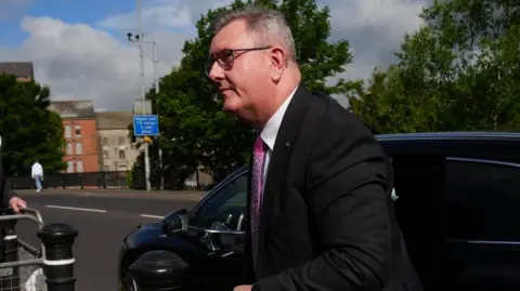A man with short greying hair exiting a black car which is on a road. He is looking to the left of the camera. He is wearing black-rimmed glasses, a black suit jacket, white shirt and pink tie. In the background other buildings, trees, street signs and a pedestrian, across the road, can be seen