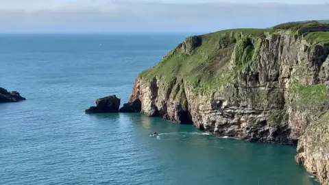 Torbay Coast and Countryside Trust Berry Head is a grassy steep-sided headland with rocky cliffs surrounded by the sea. At its end is a large rock and a small distance away from it is an island. There are two jet skis off the rocky coastline. It is a clear sky day and the blue sea is calm.