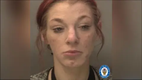 Reanne Coulson has brown hair that has been tinted red, tied back in a ponytail with wisps around her face. She has makeup smudges under her eyes and is looking down, in the photograph issued by police, and appears sad. She is wearing a top with a black neck under a jacket or jumper. She is white and aged 34.