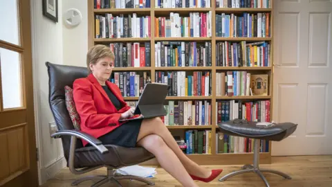 PA Media Nicola Sturgeon sitting looking an iPad in front of bookshelves. She is wearing a red jacket and a black dress.