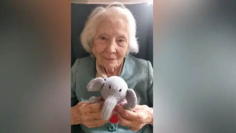 Hill Care Group Ellen Else has white hair and a grey cardigan with a peach blouse. She is sitting on a grey chair holding a knitted, stuffed toy elephant.