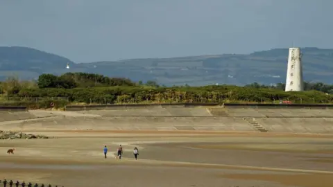 Getty Images Three people stroll with a dog on the golden sandy beach with steps going up to a white lighthouse tower surrounded by green shrubs and trees. Green hills can be seen in the background.