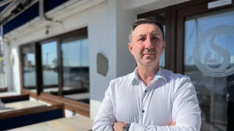 Alex La Ragione is wearing a white shirt and standing with his arms folded outside his restaurant, Sorrento.