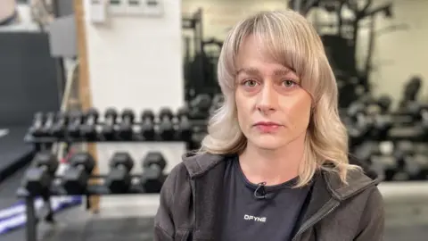 BBC Georgina Cox is sitting in a gym with dumbbells on a rack in front of a large mirror behind her. She is wearing a black t-shirt and a black hoodie over the top of it. She has blonde hair down to her shoulders and looks upset.