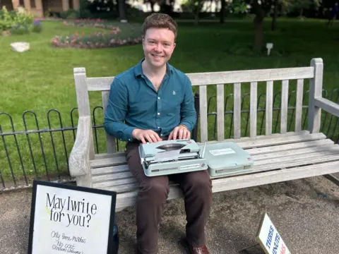 A man in brown trousers and navy shirt sits on a wooden bench, an old typewriter on his knee. Behind him are low iron railings, grass, and a flower bed.