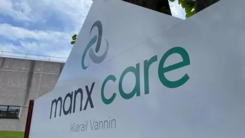 A white sign reading Manx Care in green and black writing with a Celtic swirl logo above it. 