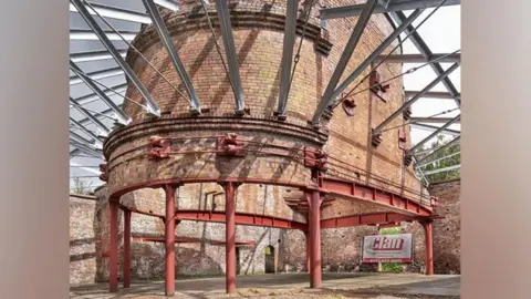 Historic England A red brick built horseshoe-shaped building is propped up on red metal columns. A frame of multiple metal beams come out of the bricks.