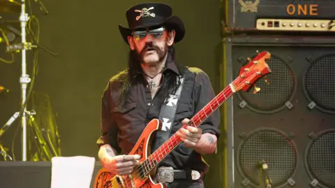 Lemmy Kilmister - wearing a black cowboy hat, an iron cross necklace, a black shirt and black trousers - holds an orange and red-coloured bass guitar while playing on stage. A large guitar amplifier and cabinet is visible behind him.  