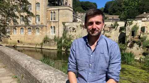 A man wearing a blue shirt sits on a low wall beside a river with a former mill building in the background.