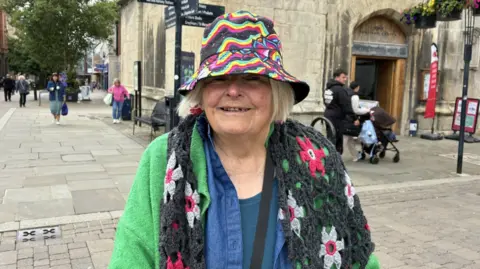 An older lady stands at The Cross in Gloucester. An old church-like tower can be seen behind her, with pedestrians behind her using the pedestrianised street.