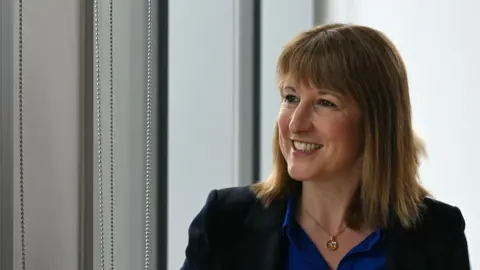 Image shows the Chancellor.
She has bobbed brown hair and is wearing a blue shirt, with a blue jacket and golden necklace. 
She is standing in front of a window looking out of it to her right. 