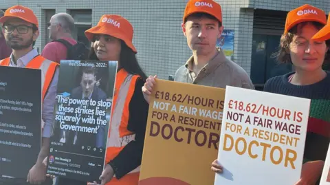 BBC Striking doctors on a picket line. they are holding signs and wearing orange hats.
